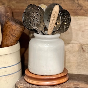 May include: A white ceramic crock holds several vintage metal butter pats with decorative cutouts. One wooden handle is clearly visible with the word BUTTER stamped into the surface. The crock sits on a round wooden base.
