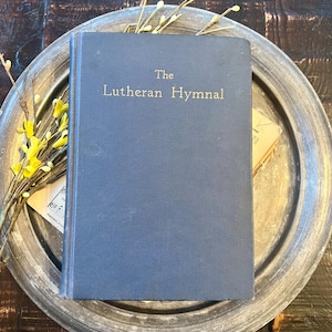 May include: A navy blue book titled "The Lutheran Hymnal" with gold lettering. The book is resting on a silver-colored metal tray, with yellow floral accents. The background is a dark wooden surface.