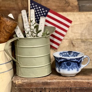 May include: A sage green metal pitcher with horizontal ridges and a handle, containing a small American flag and white folding wooden rulers. Beside it sits a white teacup featuring a detailed dark blue floral pattern and a scalloped rim.