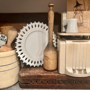 May include: A collection of vintage kitchen items including a white ceramic plate with a lace design, a wooden rolling pin, a wooden pestle, a ceramic crock with blue stripes, a white ceramic pitcher, and a vintage white kitchen scale.