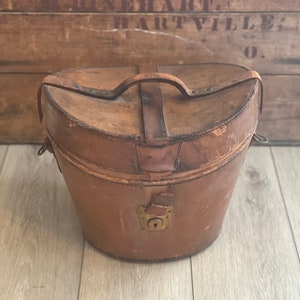 May include: A vintage brown leather hat box with a brass lock and a leather handle.