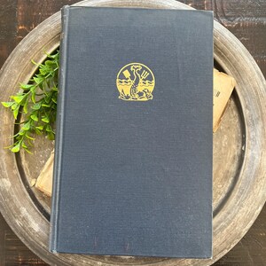 May include: A dark navy blue hardcover book with a gold circular emblem on the front cover. The emblem depicts a stylized sea creature holding a trident and a shovel, set against a background of waves. The book has a clean, solid-colored cover.