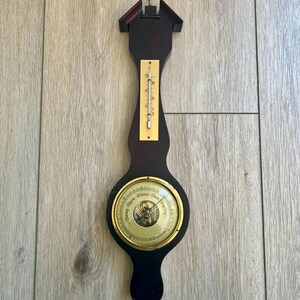 May include: A dark wood antique barometer with a thermometer and a circular dial. The dial has a gold-colored bezel and a glass cover. The thermometer is mounted on a brass plate. The barometer is hanging on a light brown wooden surface.