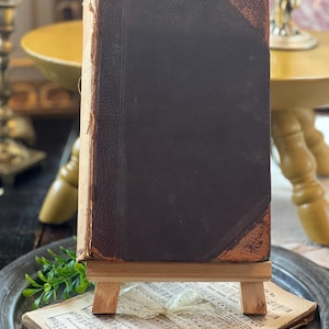 May include: A vintage, dark brown leather-bound book on a wooden easel. The book is resting on a stack of old sheet music with a white lace ribbon.