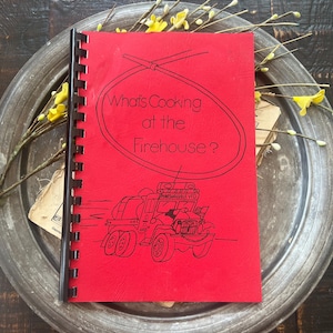 May include: A red spiral-bound cookbook titled What's Cooking at the Firehouse? featuring a black line drawing of a fire engine with the text Marshallville VFD on the front cover. The book is positioned on a round, weathered metal tray.