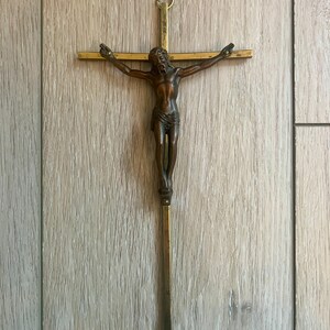 May include: A wall-mounted crucifix featuring a dark brown metallic figure of Jesus attached to a slender, polished brass cross. The cross has a simple, straight-edged design with a small circular loop at the top for hanging, showcasing a contrast between the dark bronze-toned figure and the bright, reflective gold-coloured metal of the cross structure.