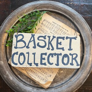 May include: A distressed wooden sign reading "BASKET COLLECTOR" in dark blue lettering. The sign is set on a metal tray with sheet music and greenery. The sign's background is off-white.