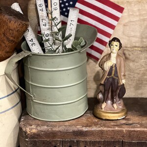 May include: A sage green metal pitcher containing a white measuring tape with numbers up to 71 inches and a small green plant. Next to it stands a small figurine of a person in a brown coat and dark trousers on a gold base, with a US flag behind.