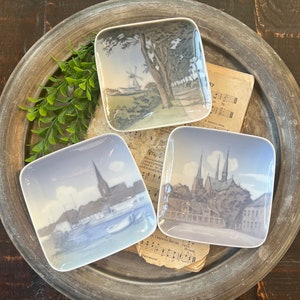 May include: Three small square ceramic dishes with blue and white painted scenes. The dishes are on a gray metal tray. The scenes are of a windmill, a city with a church, and a city with a bridge.