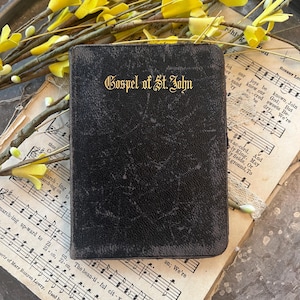 May include: A small, dark-colored book titled "Gospel of St. John" in gold lettering. The book rests on sheet music and is adorned with yellow floral sprigs. The book's cover has a textured appearance.