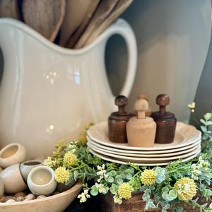 May include: A farmhouse-style display featuring a white ceramic pitcher filled with wooden spoons. Small wooden molds sit atop a stack of plates, surrounded by faux greenery and yellow flowers. A wooden bowl holds small ceramic bowls and decorative stones.