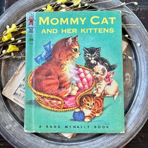 May include: A vintage children's book titled Mommy Cat and Her Kittens, featuring a green cover with an illustration of a mother cat in a basket with three kittens, one playing with a blue yarn ball. The book is a Tip-Top Elf Book by Rand McNally.