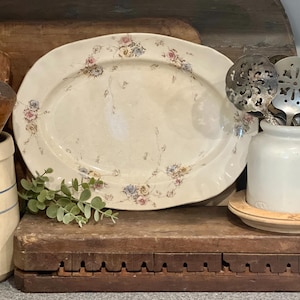 May include: An oval, off-white serving platter with floral accents in pink, blue, and yellow. A white ceramic jar holds various kitchen utensils, including slotted spoons. A small sprig of greenery rests on a wooden surface, alongside a stoneware crock.