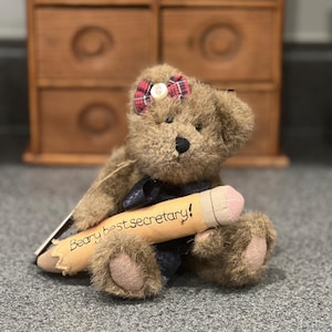 May include: A brown teddy bear wearing a red and black plaid bow and a black ribbon around its neck. The bear is holding a large pencil that says "Beary best secretary!"