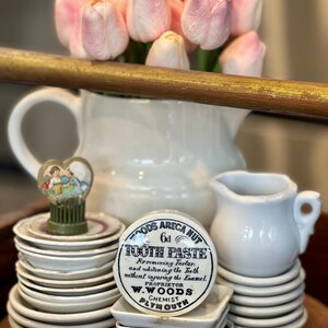 May include: A still life arrangement featuring a white pitcher with pink tulips, small white dishes, and a vintage "Tooth Paste" tin. The tin is round with black text and a white background. A small white creamer and a decorative figurine are also present.