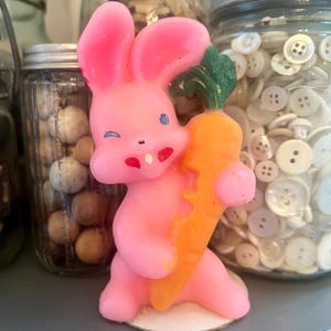 May include: A pink moulded wax candle shaped like a winking rabbit holding a large orange carrot with green leafy tops. The rabbit has painted blue eyes, a small white tooth, and red accents on its cheeks and mouth, standing on a circular base.