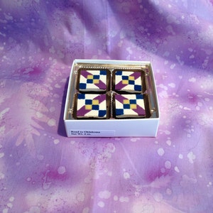 Könnte beinhalten: Eine weiße Schachtel mit einem durchsichtigen Deckel, in der sich vier kleine, quadratische Pralinen befinden. Die Pralinen sind mit einem Muster aus violetten, gelben und blauen Quadraten verziert. Die Schachtel trägt ein Etikett mit der Aufschrift "Road to Oklahoma Net Wt. 4 oz."