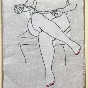 May include: An embroidered artwork depicting a seated figure in a minimalist style. The figure is rendered in black thread on a light-colored fabric, with red accents on the toes. The artwork is framed in a wooden frame.