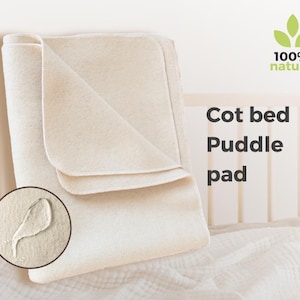 Puede incluir: Un protector de colchón para cuna de color blanquecino, doblado. El protector está hecho de un material suave y natural y lleva la etiqueta "100% natural". El texto "Cot bed Puddle pad" está en negro. Se muestra una gota de agua.