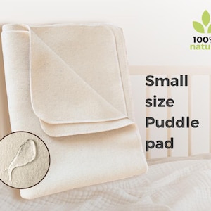 May include: A folded, off-white small size puddle pad. The image includes a close-up of a water droplet on the pad, and the text "100% natural". The pad is displayed on a white surface.