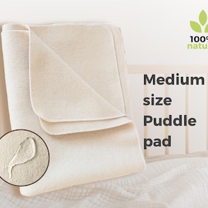 May include: A folded, cream-colored medium-size puddle pad. The pad is made of a soft, natural material and is labeled "100% natural." A close-up shows a water droplet on the pad. The text "Medium size Puddle pad" is also visible.
