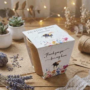 May include: A seed bomb favor with a bee and floral design. The top of the favor reads "Mr & Mrs Thomas 06/06/2027". The front of the favor reads "Thank you for bee-ing here". The favor is on a wooden surface, with other decorative items.
