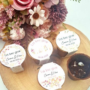 May include: Four small, brown, round pots with white labels that say "Let love grow" with a floral design and the text "Emma & Hum" and the date "09-06-2024". The pots are sitting on a wooden surface with a bouquet of pink and white flowers in the background.
