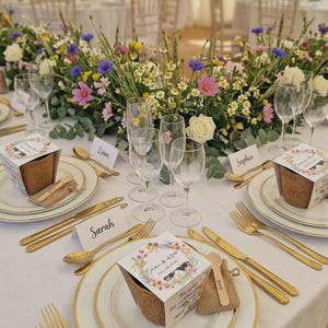 May include: A beautifully arranged wedding table featuring gold cutlery, white plates with gold edging, and floral decorations. Each place setting has a gift box and a name card. The scene suggests a formal celebration.