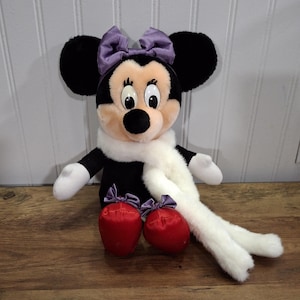 May include: A Minnie Mouse soft toy with black ears, a purple bow, and a white scarf. The doll has red shoes with purple bows. The background is a wooden surface and a white paneled wall.