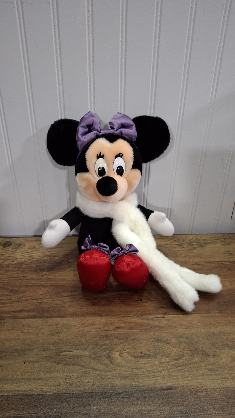 May include: A Minnie Mouse soft toy with black ears, a purple bow, and a white scarf. The doll has red shoes with purple bows. The background is a wooden surface and a white paneled wall.