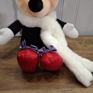 May include: A plush Minnie Mouse doll with a purple bow, black ears, and a white scarf. The doll has red, satin-like shoes with purple bows. The doll is sitting on a wooden surface.