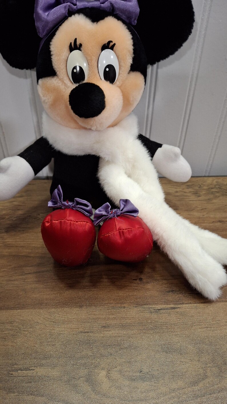 May include: A plush Minnie Mouse doll with a purple bow, black ears, and a white scarf. The doll has red, satin-like shoes with purple bows. The doll is sitting on a wooden surface.
