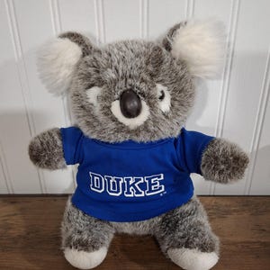 May include: A plush koala bear wearing a blue t-shirt with the word "DUKE" in white block letters. The koala has gray fur, white ears, and white paws. The background is a white paneled wall.