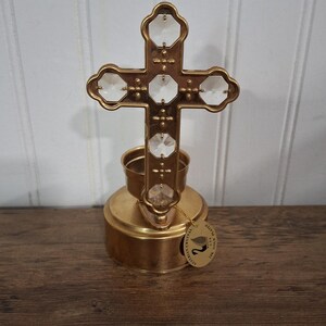 May include: A gold-colored cross with clear crystal accents, mounted on a matching base. The cross features a decorative design and is likely a religious item. The base has a circular shape. A tag hangs from the base.