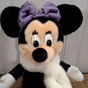 May include: A Minnie Mouse soft toy with black ears, a black and white body, and a light pink face. She has a purple bow, a white scarf, and red shoes with purple bows. A classic Disney character.