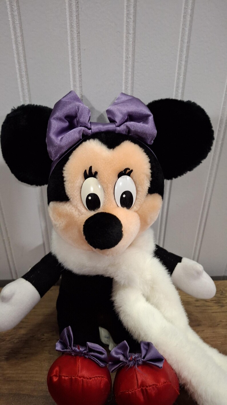 May include: A Minnie Mouse soft toy with black ears, a black and white body, and a light pink face. She has a purple bow, a white scarf, and red shoes with purple bows. A classic Disney character.