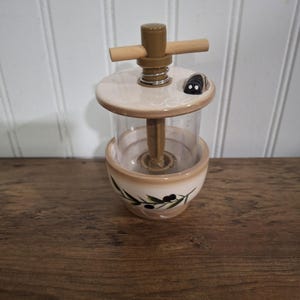 May include: A beige and clear olive oil dispenser with a wooden handle and a small black and white bug-shaped accent. The ceramic base features a green olive branch design. The dispenser sits on a wooden surface.