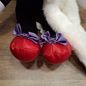 May include: Close-up of a plush toy's feet. The feet are red, round, and adorned with purple satin bows. The toy's legs are black, and the body is white and black. The background is a wooden surface.