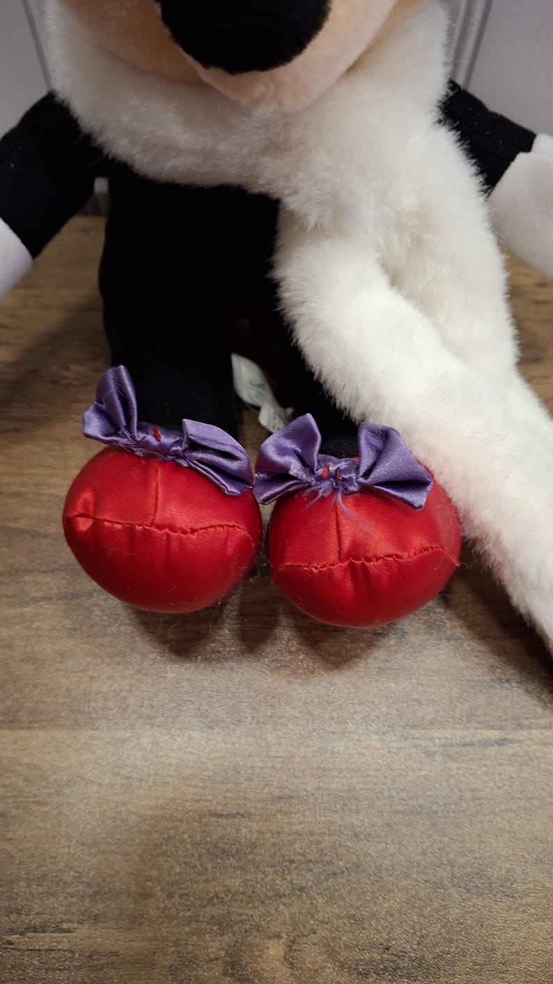 May include: Close-up of a plush toy's feet. The feet are red, round, and adorned with purple satin bows. The toy's legs are black, and the body is white and black. The background is a wooden surface.