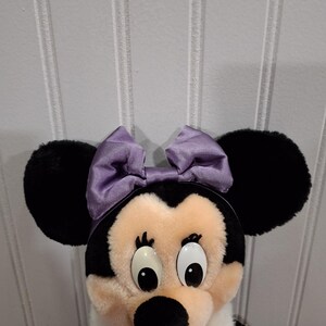 May include: A Minnie Mouse soft toy featuring black ears, a lavender bow, and a white scarf. The doll has a black nose, large eyes, and a black and white body. The backdrop is a white panelled wall.