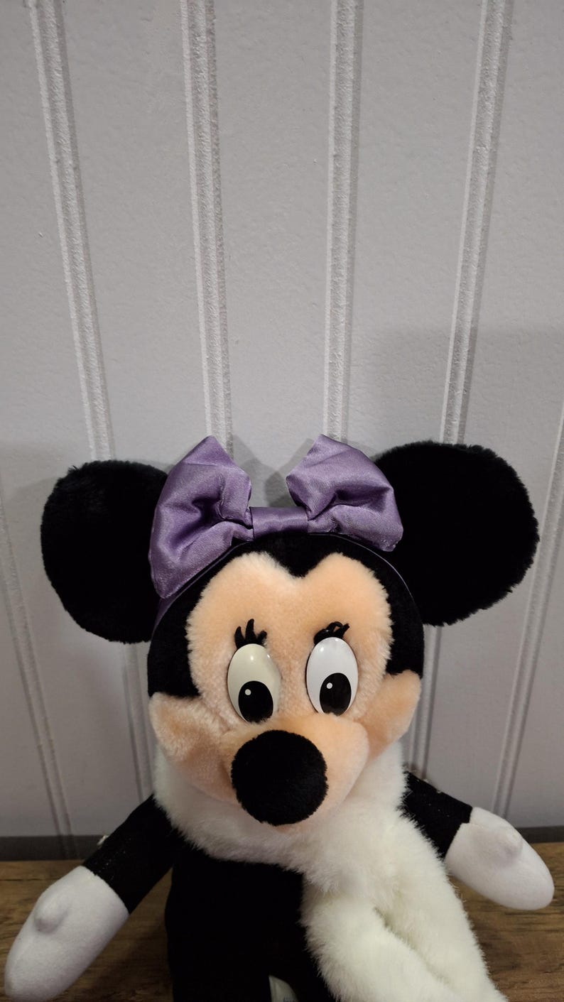 May include: A Minnie Mouse soft toy featuring black ears, a lavender bow, and a white scarf. The doll has a black nose, large eyes, and a black and white body. The backdrop is a white panelled wall.