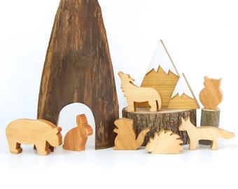 Könnte beinhalten: Holzspielzeug-Set mit verschiedenen Tieren, darunter ein Wolf, eine Eule, ein Fuchs, ein Kaninchen und ein Igel. Das Set enthält auch einen Holzberg und einen Baumstumpf. Die Spielzeuge sind alle aus Naturholz gefertigt und in einer hellbraunen Farbe lackiert.