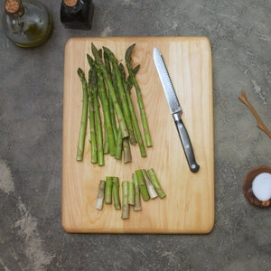 May include: A wooden cutting board with a bunch of green asparagus spears, a silver knife, and a small bowl of salt.