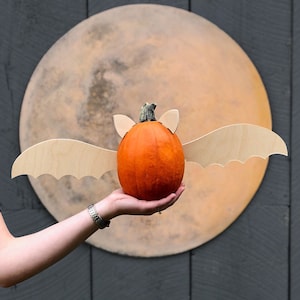 May include: A small orange pumpkin with wooden bat wings attached to it. The pumpkin is being held in front of a large, round, brown and white painted moon.