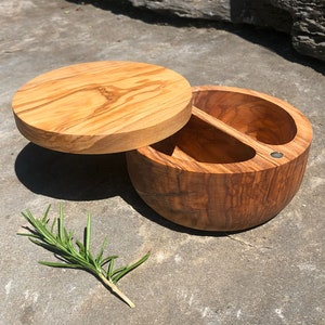 May include: A wooden salt cellar with a lid, featuring a two-compartment design. The natural wood grain is visible, and a sprig of rosemary lies nearby. The lid is slightly ajar, revealing the interior. The container is approximately 4 inches in diameter.