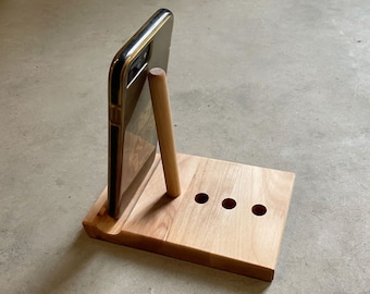 May include: A light-colored wooden phone stand with a slot for a phone and three small holes. A phone with a gold-colored frame is in the stand, supported by a wooden dowel. The stand is made of light-colored wood.
