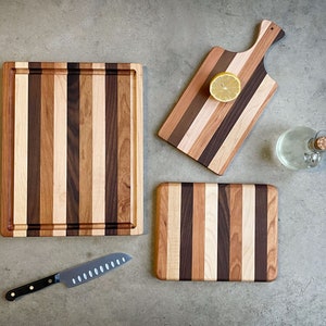 May include: Three wooden cutting boards with a striped pattern of different wood types. The boards are all rectangular and have rounded corners. The largest board is 16 inches long and 12 inches wide. The middle board is 12 inches long and 9 inches wide. The smallest board is 8 inches long and 6 inches wide.