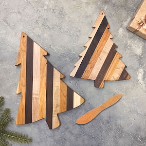 May include: Two wooden Christmas tree shaped cutting boards with a striped pattern in shades of brown, black, and light wood. A wooden butter knife is included.