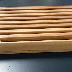 May include: A rectangular wooden breadboard with a slatted top. The breadboard is made of light and dark brown wood, with a box-like frame. The slats are evenly spaced and run horizontally across the top. The wood grain is visible.
