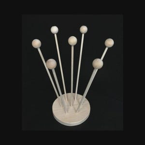 May include: A wooden display stand with a circular base and several thin, vertical wooden rods, each topped with a small, round wooden ball. The stand is made of light-colored wood and is set against a black background.